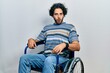 Handsome hispanic man sitting on wheelchair in shock face, looking skeptical and sarcastic, surprised with open mouth