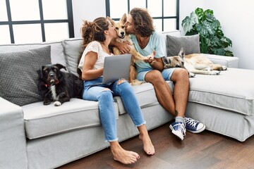 Man and woman couple using laptop sitting on sofa kissing dog at home