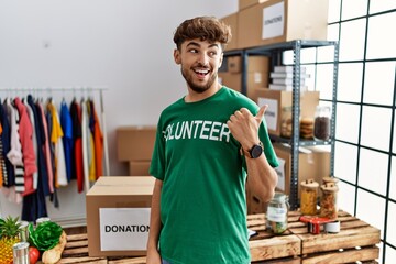 Poster - Young arab man wearing volunteer t shirt at donations stand smiling with happy face looking and pointing to the side with thumb up.