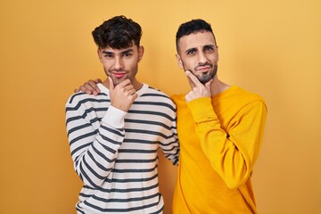 Poster - Young hispanic gay couple standing over yellow background looking confident at the camera smiling with crossed arms and hand raised on chin. thinking positive.