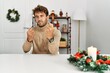 Young handsome man with beard sitting on the table by christmas decoration ready to fight with fist defense gesture, angry and upset face, afraid of problem