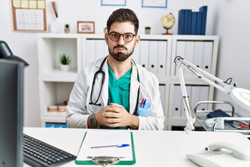 Sticker - Young man with beard wearing doctor uniform and stethoscope at the clinic puffing cheeks with funny face. mouth inflated with air, crazy expression.