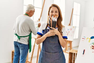 Wall Mural - Two middle age artists smiling happy painting at art studio. Woman standing covering face with paintbrushes.