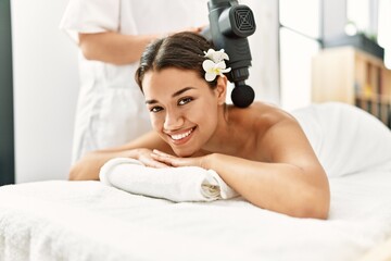 Poster - Young latin woman relaxed having back massage with percussion pistol at beauty center