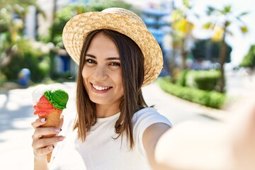 Young beautiful woman smiling happy outdoors on a sunny day wearing a summer hat and eating ice cream taking a selfie picture