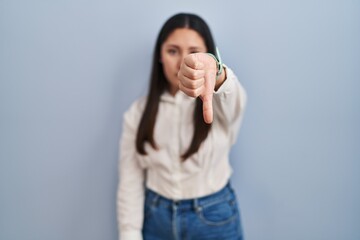 Poster - Young latin woman standing over blue background looking unhappy and angry showing rejection and negative with thumbs down gesture. bad expression.