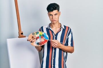 Sticker - Young hispanic man holding painter palette and paintbrush standing close to canvas depressed and worry for distress, crying angry and afraid. sad expression.