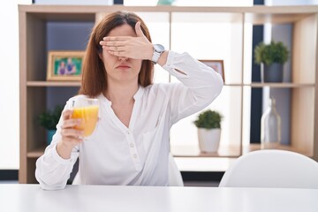 Poster - Brunette woman drinking glass of orange juice covering eyes with hand, looking serious and sad. sightless, hiding and rejection concept