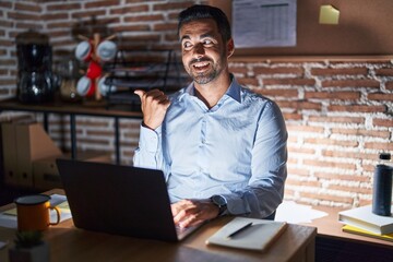 Canvas Print - Hispanic man with beard working at the office at night smiling with happy face looking and pointing to the side with thumb up.