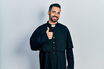 Canvas Print - Handsome hispanic man with beard wearing catholic priest robe doing happy thumbs up gesture with hand. approving expression looking at the camera showing success.