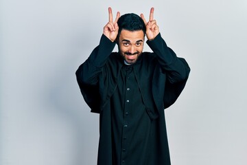 Canvas Print - Handsome hispanic man with beard wearing catholic priest robe posing funny and crazy with fingers on head as bunny ears, smiling cheerful