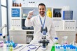 © Krakenimages.com - Young hispanic man with beard working at scientist laboratory smiling and confident gesturing with hand doing small size sign with fingers looking and the camera. measure concept.
