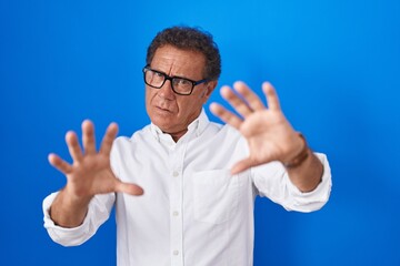 Wall Mural - Middle age hispanic man standing over blue background afraid and terrified with fear expression stop gesture with hands, shouting in shock. panic concept.