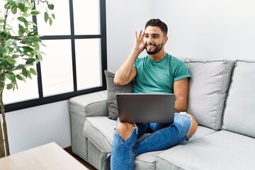 Sticker - Young handsome man with beard using computer laptop sitting on the sofa at home smiling with hand over ear listening an hearing to rumor or gossip. deafness concept.