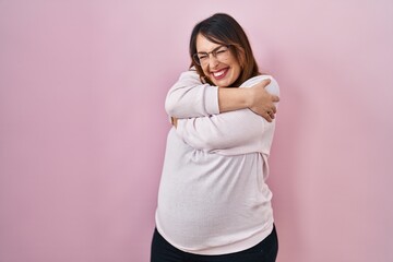 Poster - Pregnant woman standing over pink background hugging oneself happy and positive, smiling confident. self love and self care