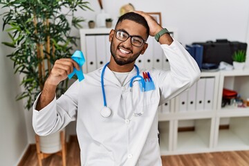 Poster - Young indian doctor holding blue ribbon stressed and frustrated with hand on head, surprised and angry face