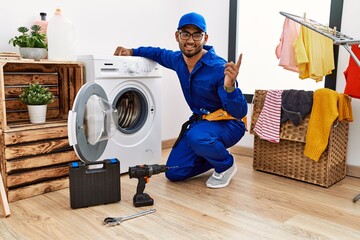 Wall Mural - Young indian technician working on washing machine with a big smile on face, pointing with hand finger to the side looking at the camera.