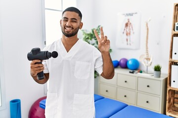 Canvas Print - Young indian physiotherapist holding therapy massage gun at wellness center smiling looking to the camera showing fingers doing victory sign. number two.