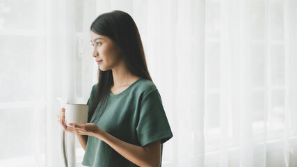 Poster - Young asian woman standing beside window and holding mug in bedroom at home, She drinking milk after wake up in the morning