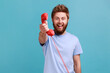 © khosrork - Portrait of smiling positive bearded man holding in hands red retro telephone, waiting for your call, looking at camera with excited expression. Indoor studio shot isolated on blue background.