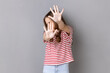 Portrait of scared little dark haired girl wearing striped T-shirt gesturing stop with palms and looking turning away, being frighten. Indoor studio shot isolated on gray background.