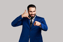 Delighted Bearded Man Standing With Telephone Hand Gesture And Smiling To Camera, Flirting Offering To Contact By Phone, Pointing Finger To Camera. Indoor Studio Shot Isolated On Gray Background.