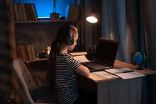 Little Girl In Wireless Headset Using Laptop, Studying Online At Home At Night, Interested Student Typing On Keyboard Looking At Pc Screen, Watching Webinar, Online Course, Doing Homework