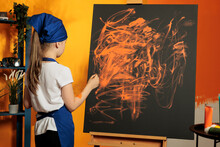 Little Girl Using Orange Aquarelle To Paint Artwork On Canvas With Easel, Doing Artistic Practice To Create Masterpiece Design With Watercolor Palette And Paintbrush. Painting Creation With Wet Dye.
