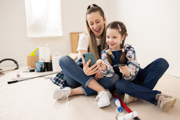 Beautiful blonde woman and girl are sitting on floor in apartment under renovation. Child points to a photo on phone of dream music center, which the girl wants to put in new room.