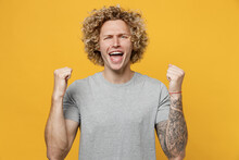 Young Overjoyed Exultant Fun Caucasian Man 20s He Wear Grey T-shirt Doing Winner Gesture Celebrate Clenching Fists Say Yes Isolated On Plain Yellow Backround Studio Portrait. People Lifestyle Concept.