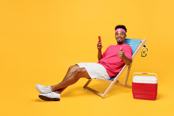 Wall Mural - Full body young happy satisfied man he wear pink t-shirt bandana near hotel pool hold in hand use mobile cell phone show thumb up isolated on plain yellow background. Summer vacation sea rest concept.