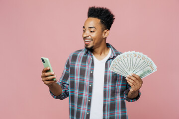Wall Mural - Young smiling fun happy man of African American ethnicity 20s wear blue shirt hold in hand use mobile cell phone fan of cash money in dollar banknotes isolated on plain pastel light pink background.