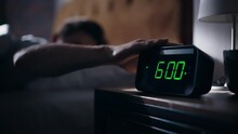 Man Wakes Up, Turns Off Alarm Clock With Frustration. Early Rising Productive Man Ready Start A Day Full Of New Problems. Focus On The Clock Showing Six A.M. Bedside Nightstand Bedroom Apartment