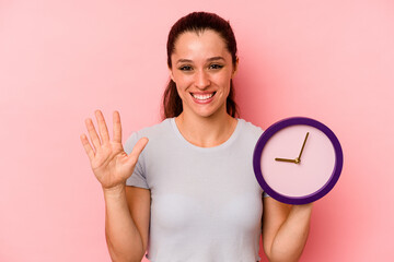 Wall Mural - Young caucasian woman holding a clock isolated on pink background smiling cheerful showing number five with fingers.