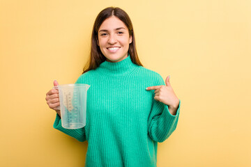 Wall Mural - Young caucasian woman holding measuring jug isolated on yellow background person pointing by hand to a shirt copy space, proud and confident