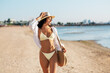 © Syda Productions - people, summer holidays and leisure concept - happy young woman in bikini swimsuit, white shirt and straw hat with bag walking along beach