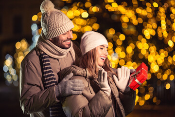 Poster - winter holidays and people concept - happy smiling couple with gift over christmas lights in evening