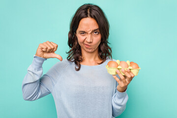 Wall Mural - Young hispanic woman holding eggs isolated on blue background showing a dislike gesture, thumbs down. Disagreement concept.