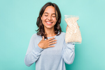 Wall Mural - Young hispanic woman holding a bag of oats isolated on blue background laughs out loudly keeping hand on chest.