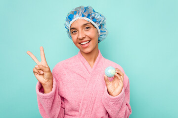 Wall Mural - Young hispanic woman holding bathtub ball isolated on blue background joyful and carefree showing a peace symbol with fingers.