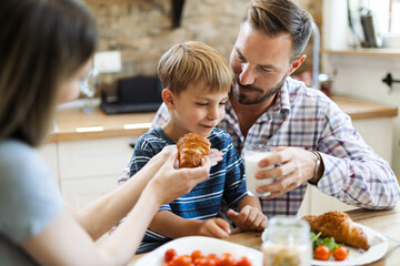 Wall Mural - Young happy parents feeding their son during breakfast time in the kitchen. Focus is on man