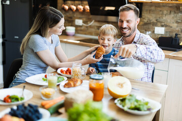 Wall Mural - Young happy parents feeding their son with croissant during breakfast time in the kitchen