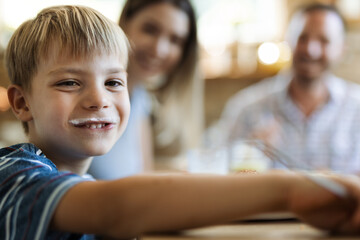 Wall Mural - Happy little boy having yogurt mustache at home and looking at camera. There are people in the background