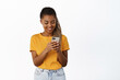 © Cookie Studio - Smiling african american girl chatting on mobile messanger, using smartphone app, standing over white background