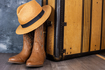 Leather cowgirl boots with straw hat pattern and an old suitcase stand on the floor against  gray wall.