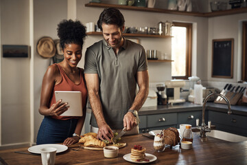 Wall Mural - Couple cooking breakfast with a tablet while searching for online recipes and videos on the internet. Happy interracial lovers preparing a healthy meal in their home kitchen while browsing online