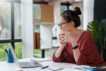 Wall Mural - Beautiful Asian woman relaxing with coffee at her office desk and looking at her laptop computer.