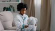 © Yuliia - Serious african american kid girl sitting in room on couch in medical uniform pretending to be doctor little child playing veterinarian heals toy patient holding stethoscope listening sick teddy bear