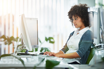 Poster - Young female designer working at her computer by a desk in a modern office. Creative business woman in design typing advertising and marketing designs on her pc keyboard at the workplace