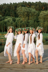 Wall Mural - Young girls in white dresses and with braided wreaths pose by the lake. Celebration of summer Kupala